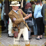 Un homme vêtu comme un explorateur du 19e siècle, arborant également un pistolet et des lunettes de style steampunk, qui utilise un appareil photo moderne au milieu d’un marché.